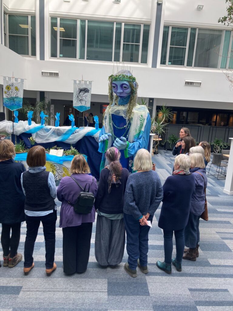 Photo of people standing in a circle singing to a large blue puppet